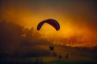 A solo person on a glider flying low to the ground surrounded by clouds and a dark orange sky getting ready to land.