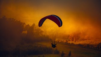 A solo person on a glider flying low to the ground surrounded by clouds and a dark orange sky getting ready to land.