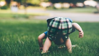 The little boy in plaid shorts is playing in the green grass bent over, exploring the ground with curiosity.