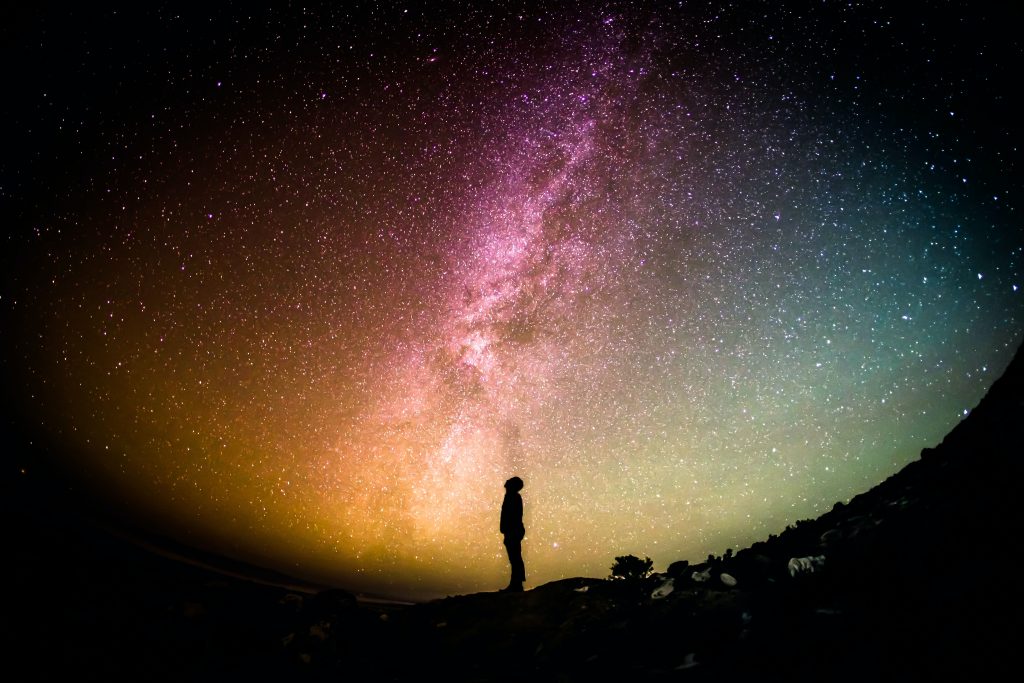 Man standing sideways in black silhouette looking up at the sky in colors of muted pink, yellow, orange, blue and green. The image of the sky is a half circle with trees and land in a half circle below his feet.