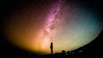 Man standing sideways in silhouette in front of a large image of the earth/universe in colors of muted pink, yellow, orange, blue and green. contemplating life