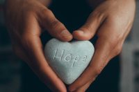 A dark skin man holding his hands in a heart while holding a gray rock with the words hope inscribed in the rock