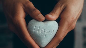 A dark skin man holding his hands in a heart while holding a gray rock with the words hope inscribed in the rock