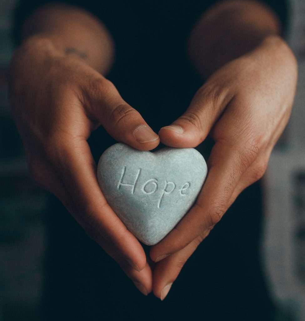 A dark skin man holding his hands in a heart while holding a gray rock with the words hope inscribed in the rock