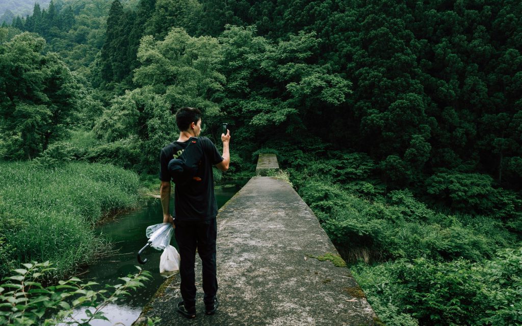Asian man in black jeans and black tee-shirt with a dark backpack standing on a walkway taking a picture with a smart phone of the green trees and forest in front of him.