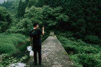 Asian man in black jeans and black tee-shirt holding an umbrella, with a dark backpack standing on a walkway taking a picture with a smart phone of the green trees and forest in front of him.